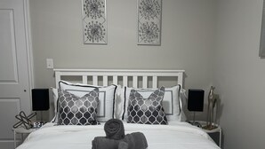 Una cama bien hecha con cabecera blanca, almohadas estampadas en gris y una toalla gris. Hay dos altavoces en las mesitas de noche y un reloj en una de ellas.