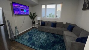 Una sala de estar con un televisor de pantalla plana grande montado en la pared, un sofá gris con cojines azules, un tapete azul estampado y una ventana con persianas.