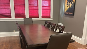 Ein Esszimmer mit Holztisch und -stühlen, großen Fenstern mit roten Vorhängen und einem Poster an der Wand.