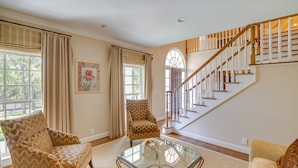 A living room with a glass coffee table, two patterned armchairs, a beige sofa, a staircase with wooden railings, and a large window with curtains. - Pool, Hot Tub & Outdoor Theater: Franklin Retreat (Franklin)