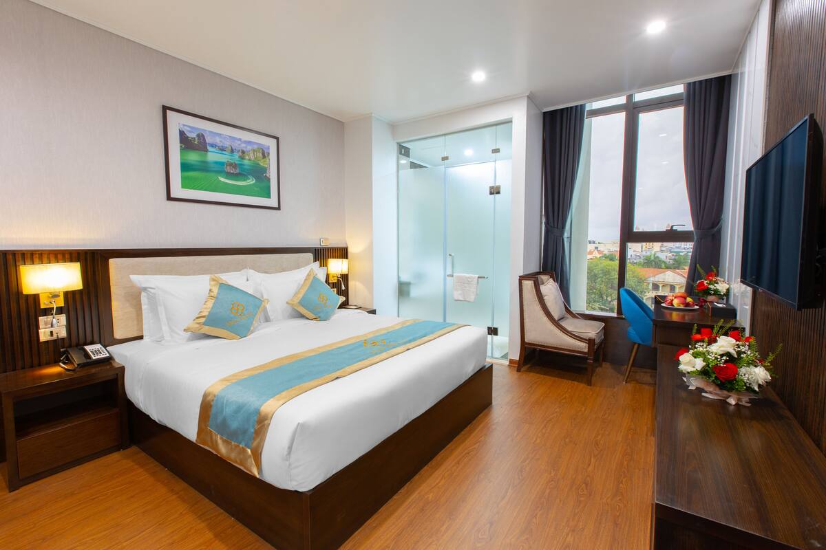 Hotelkamer met een groot bed, een bureau met stoel, een douchecabine van glas en uitzicht op de stad.