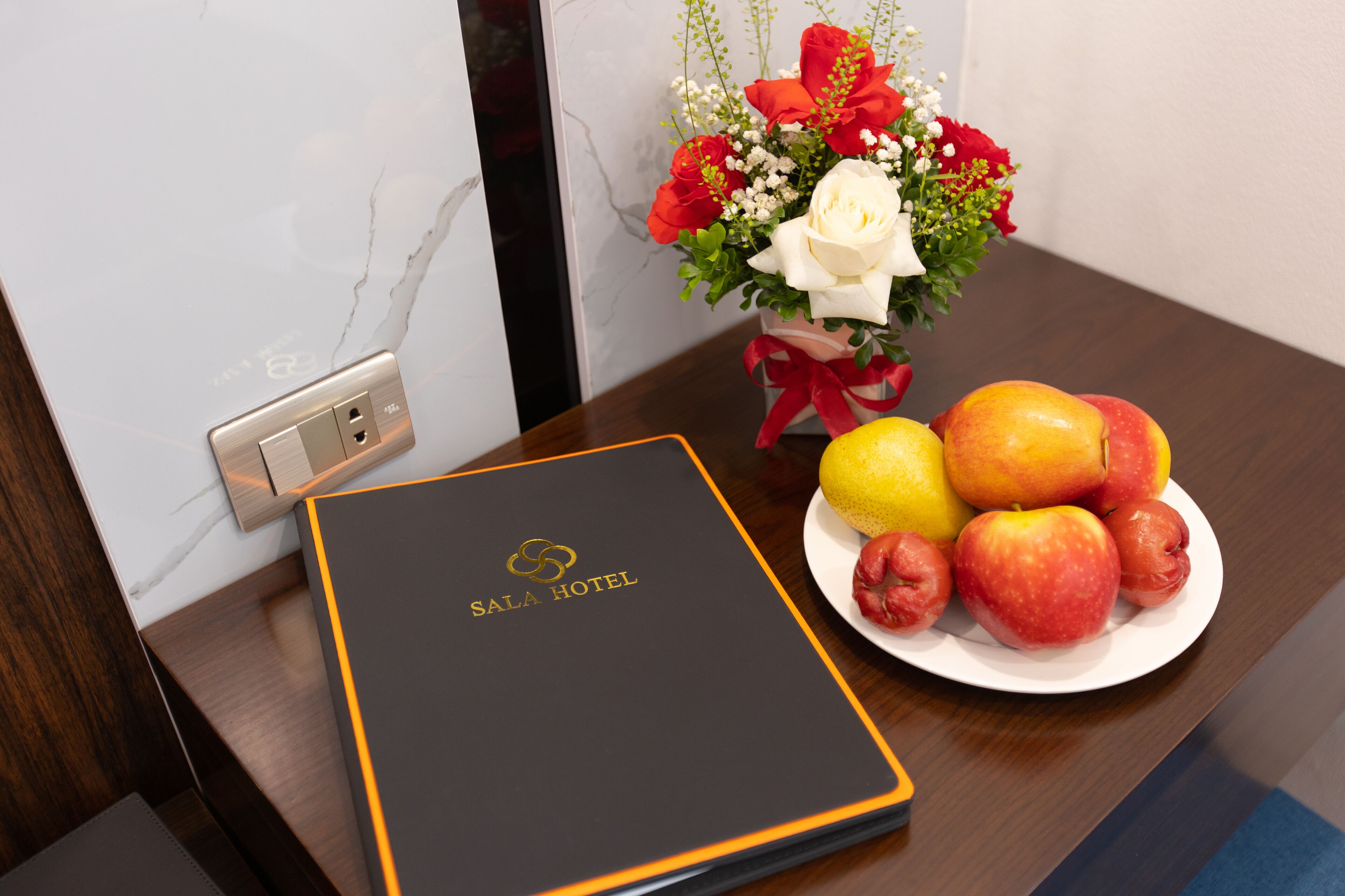 A room with a wooden table, a white plate with fruits, a vase with flowers, and a black and gold folder labeled "SALA HOTEL".