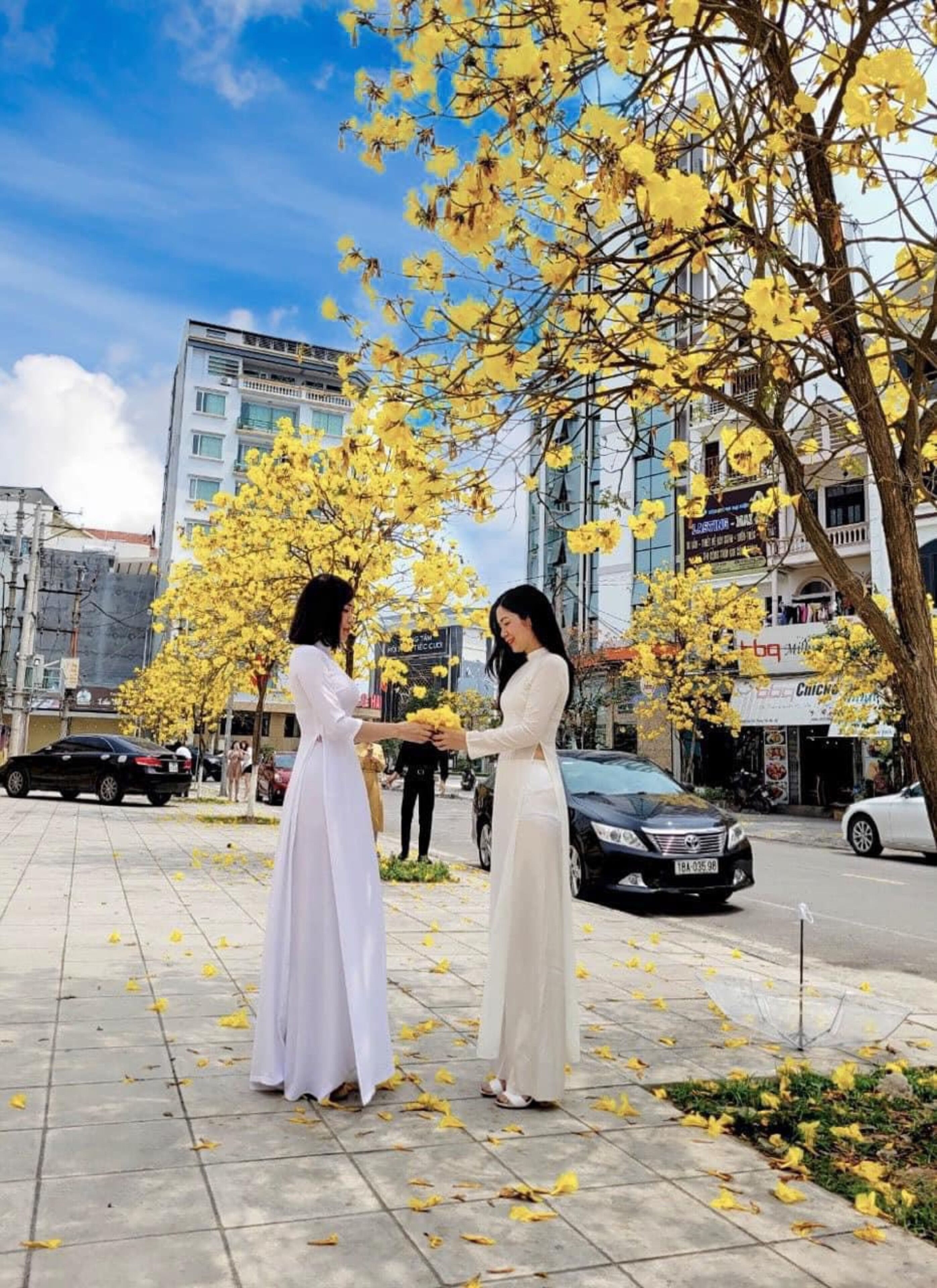 Two women in white dresses standing on a sidewalk with yellow flowers and buildings in the background.