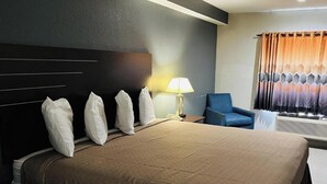 A hotel room with a large bed, a bedside table with a lamp, a blue armchair, and a window with curtains. - Regency Inn & Suites Houston East I 10 (Houston)