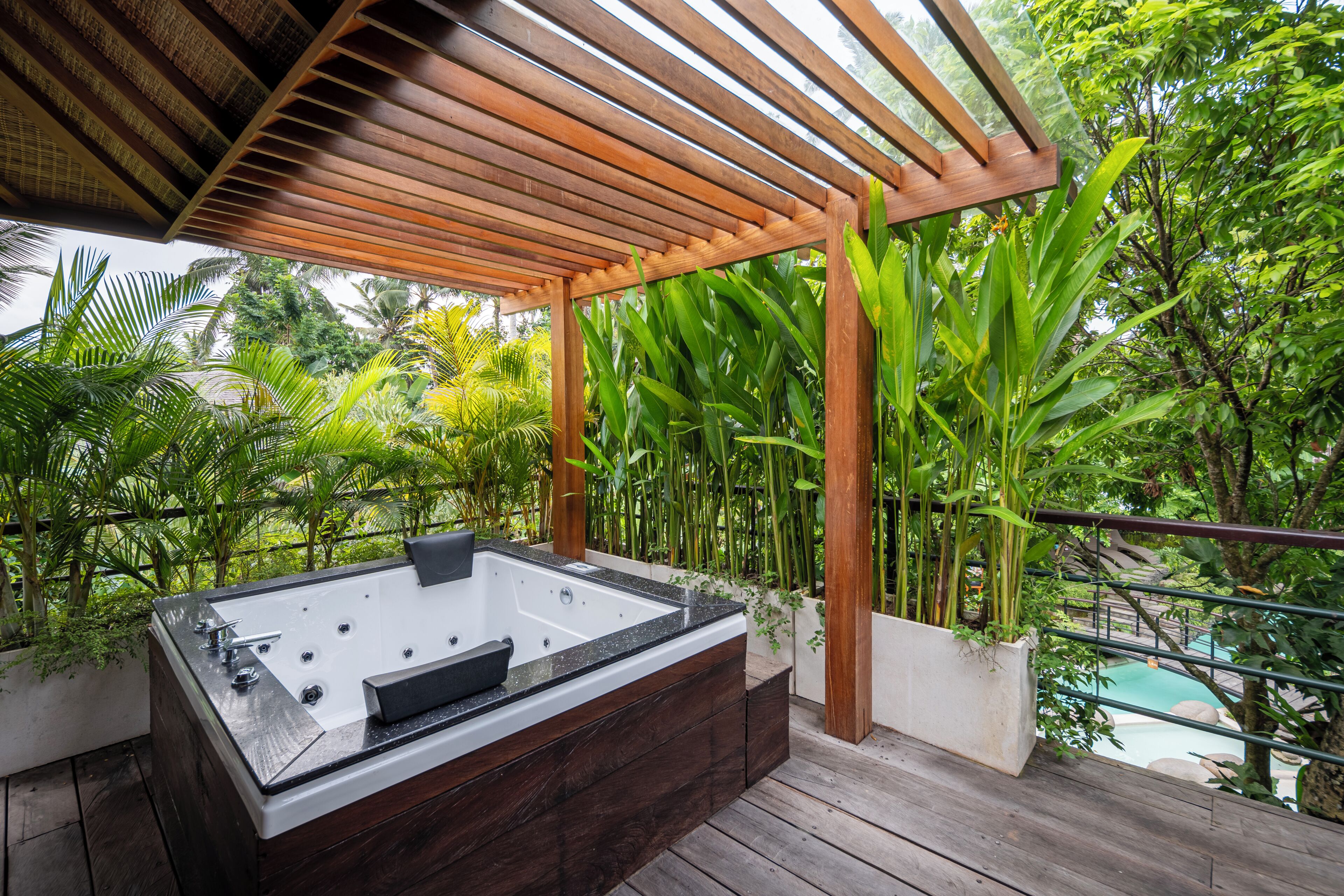 Um deck de madeira com hidromassagem, cercado por vegetação exuberante e vista para uma piscina.