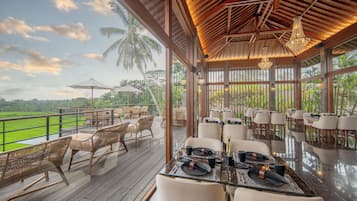 A restaurant with a wooden deck overlooking a green landscape, featuring wicker chairs and a glass dining area.