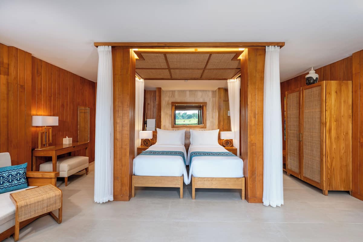 A room with two beds, wooden walls, a view through a window, and a white curtain.