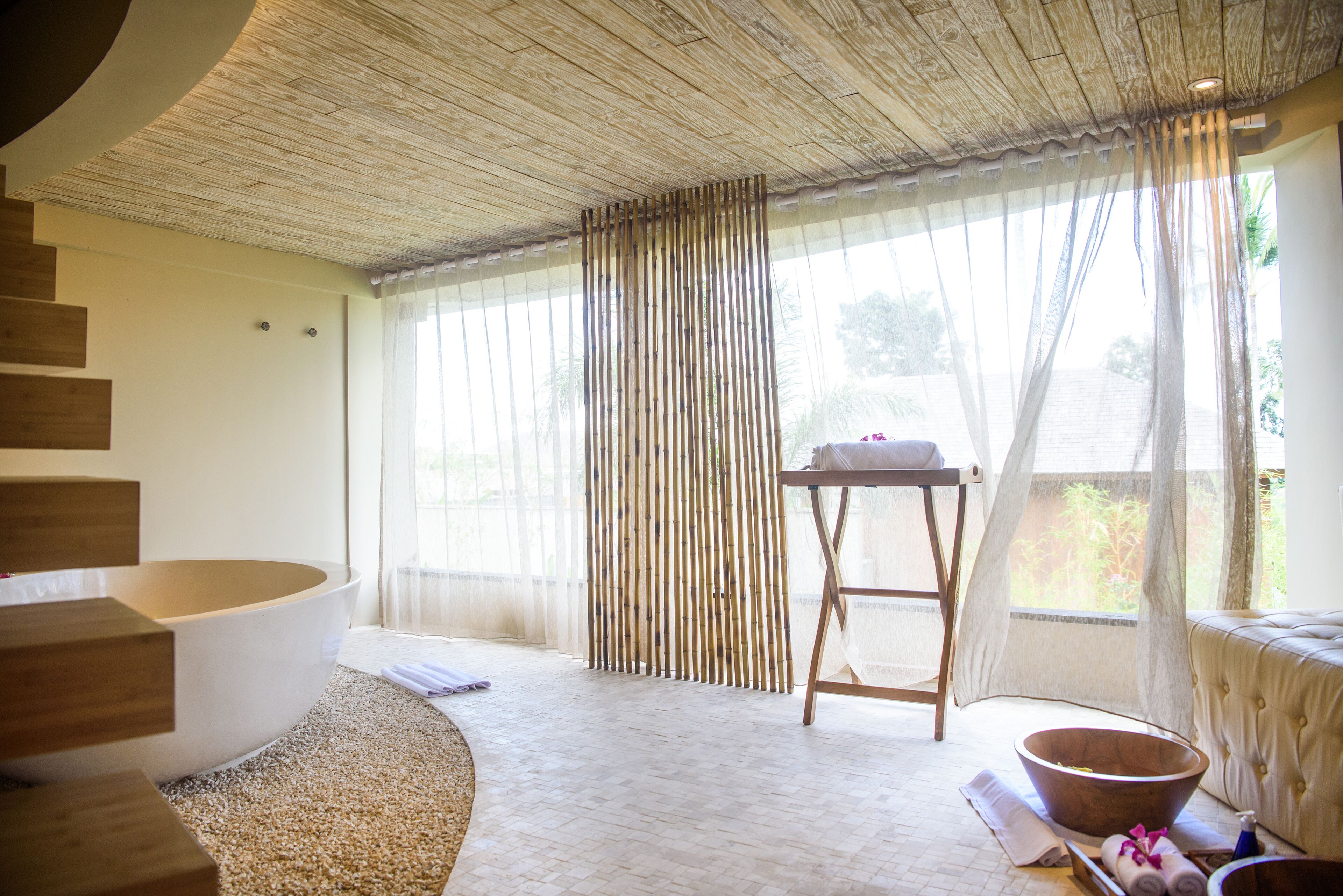 A spa room with a large bathtub, wooden flooring, a wooden staircase, and a large window with sheer curtains.