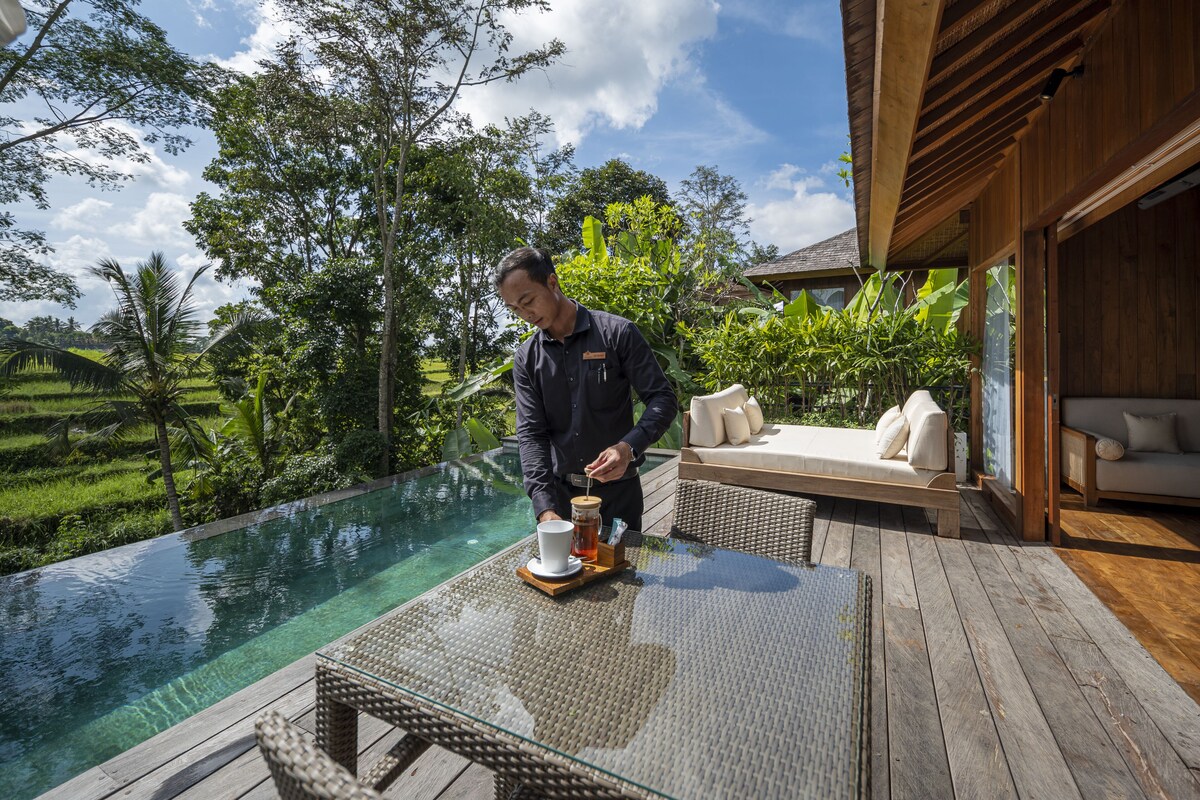 Un hombre sirviendo una bebida en una mesa junto a una piscina infinita con terraza de madera, rodeado de exuberante vegetación y un edificio moderno.