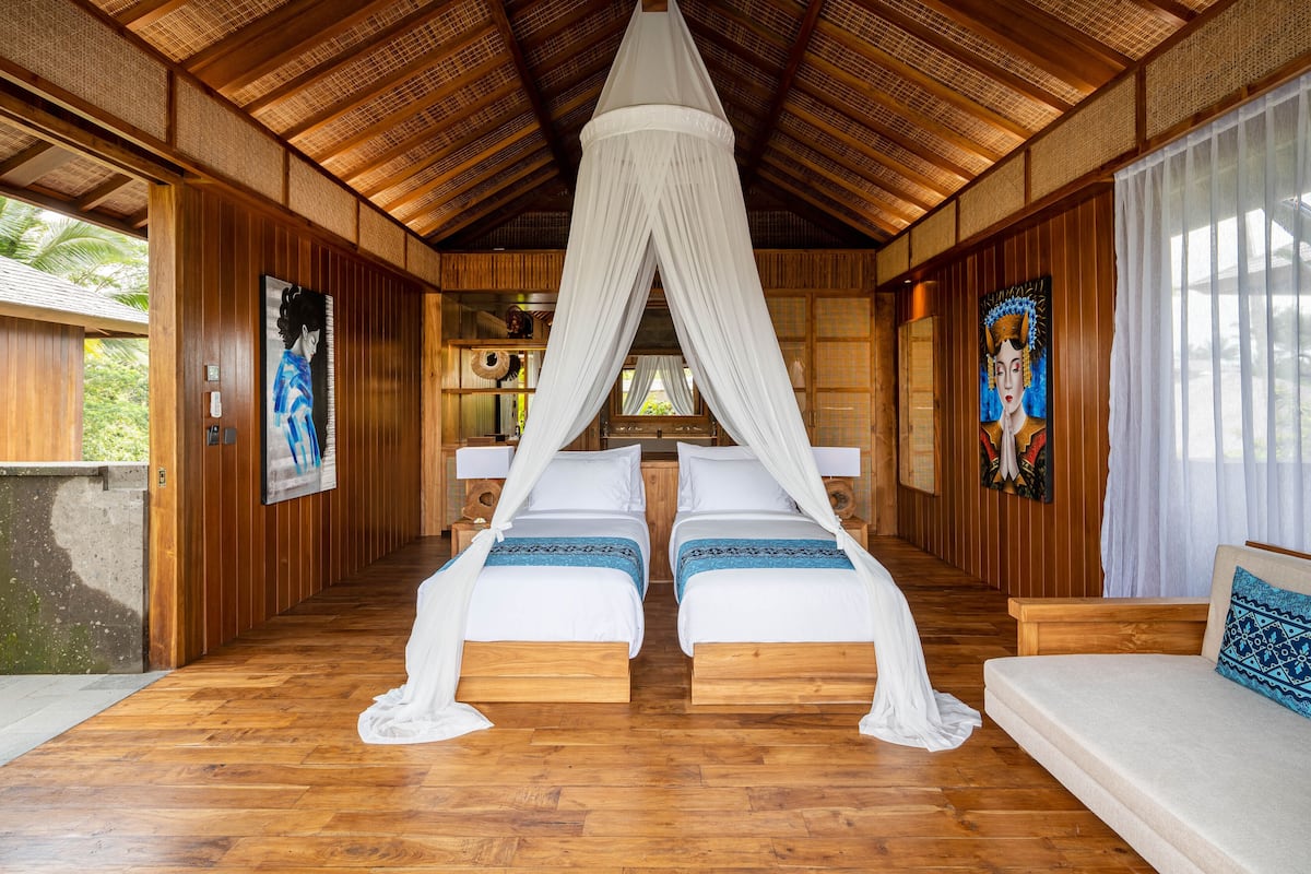 A room with two beds, a canopy bed, wooden walls, and a view of greenery outside.
