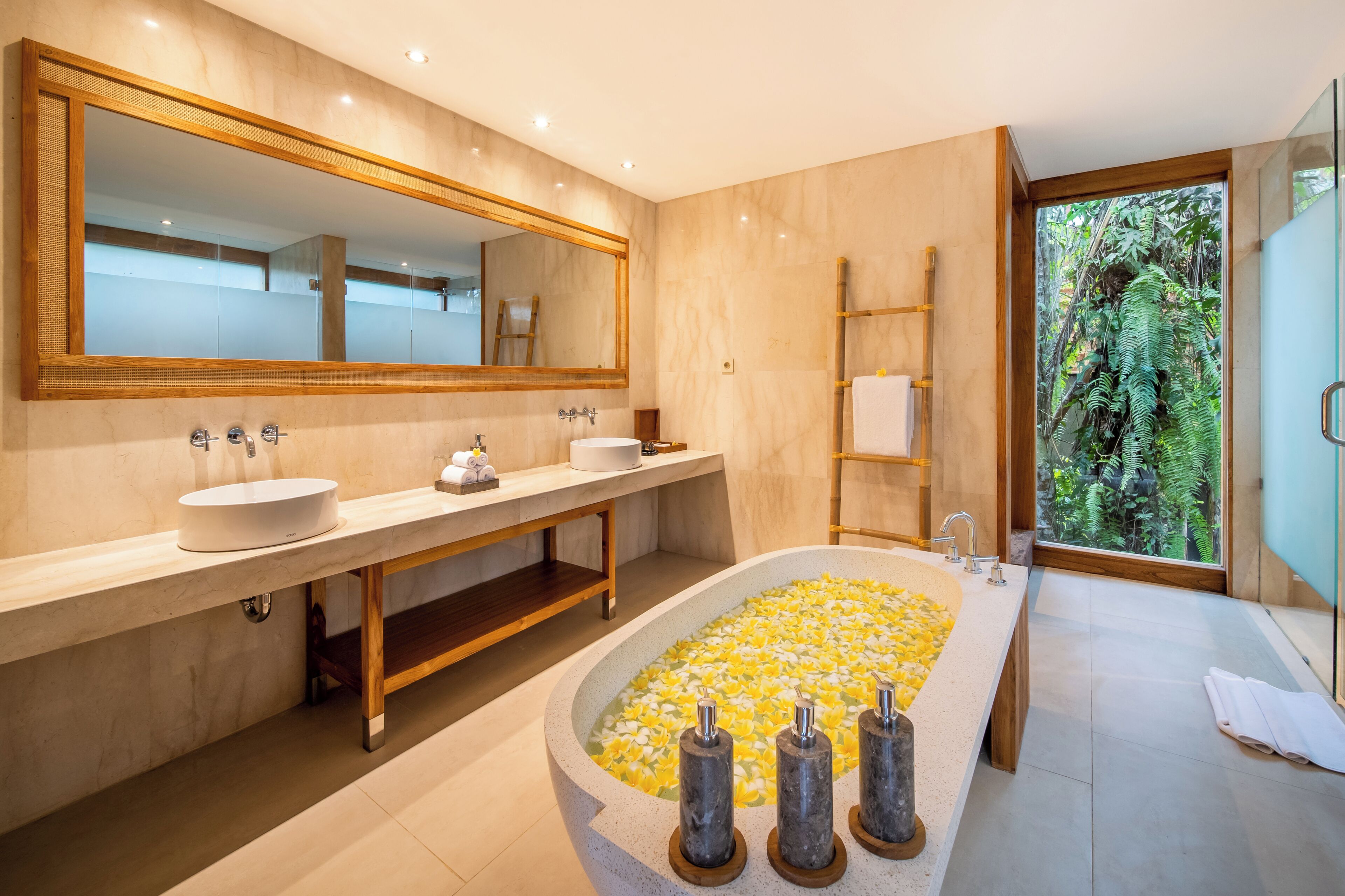 A modern bathroom with a large oval bathtub, two sinks, a large mirror, and a view of greenery through a window.