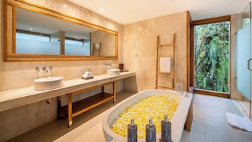 Ein modernes Badezimmer mit einer großen, ovalen Badewanne, zwei Waschbecken, einem großen Spiegel und Blick auf Grün durch ein Fenster.