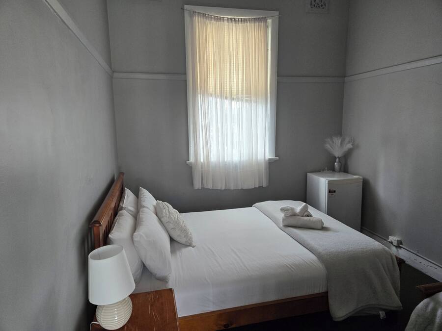 Un dormitorio con cama, mesita de noche, lámpara, ventana con cortinas y un mueble bajo.
