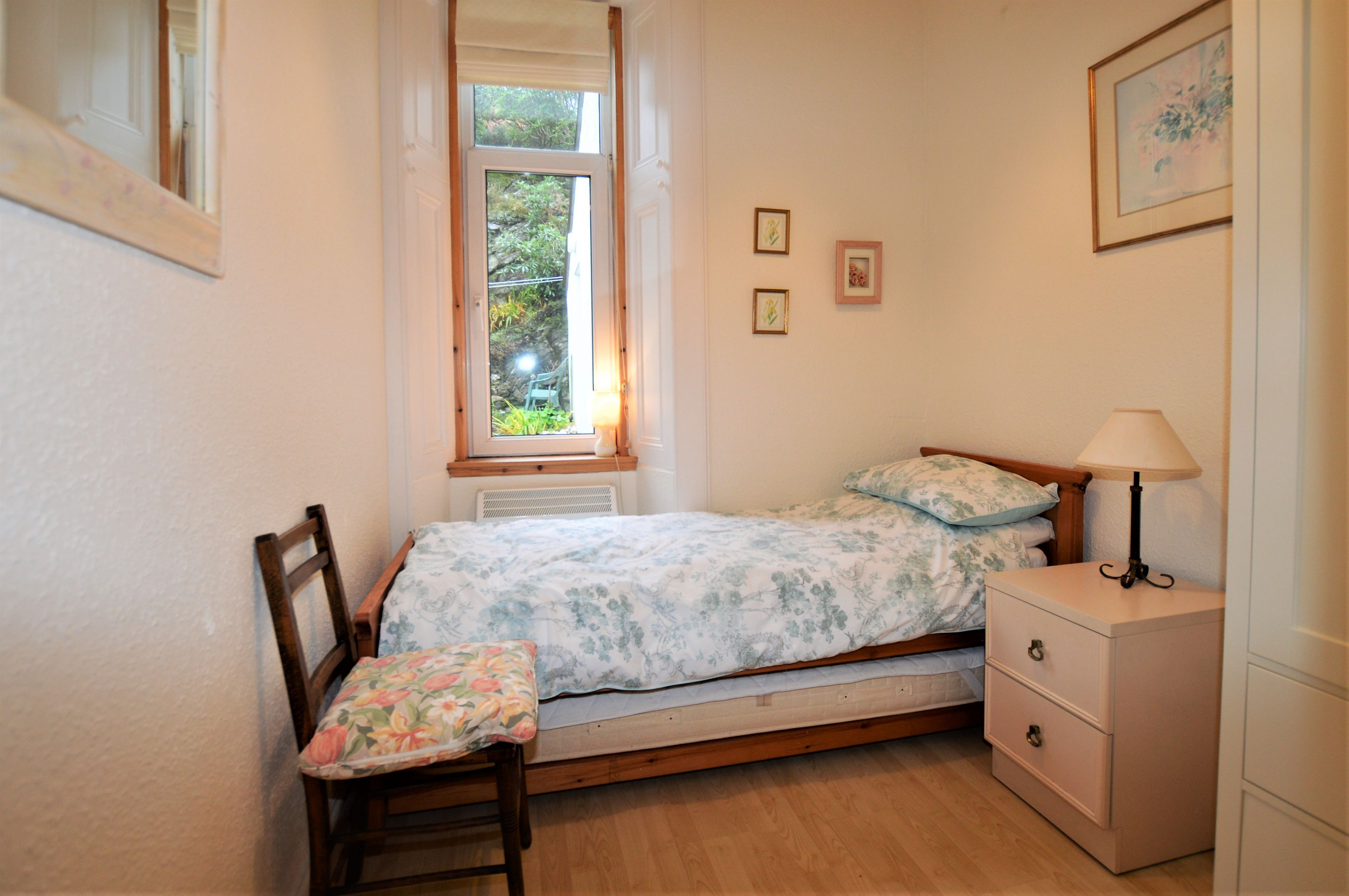 A small bedroom with a single bed, a chair, a nightstand, and a window with a view of greenery.