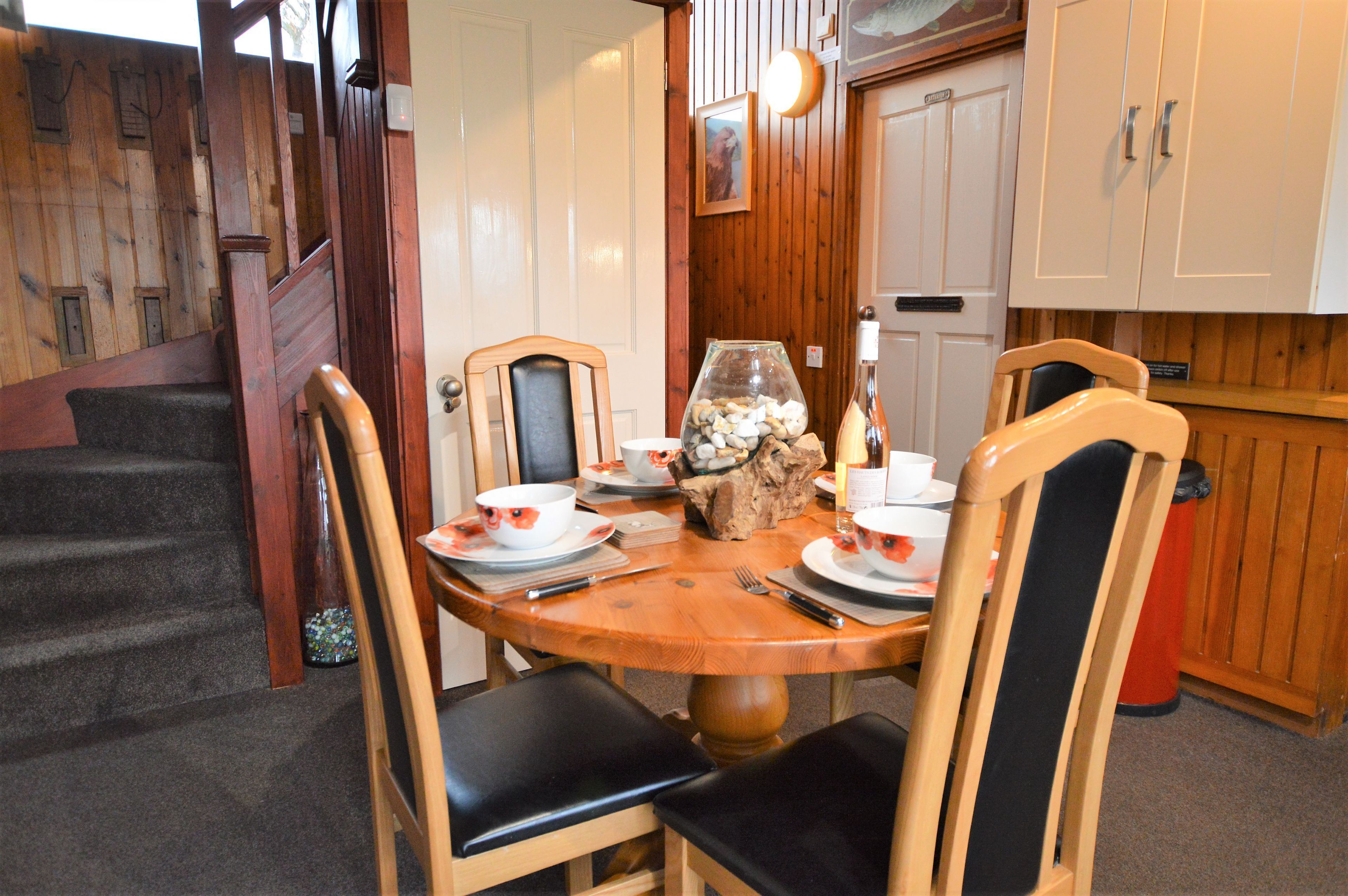 Een eetruimte met een houten tafel gedekt voor vier personen, omringd door stoelen met zwarte kussens. Op tafel staan een vaas met stenen en een kaars.