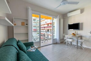A modern living room with a green sofa, a dining table with chairs, a TV mounted on the wall, and a balcony with a view of a residential building.