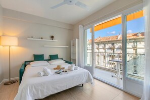 A hotel room with a bed, a standing lamp, a ceiling fan, a balcony with a view of buildings, and a window with curtains.