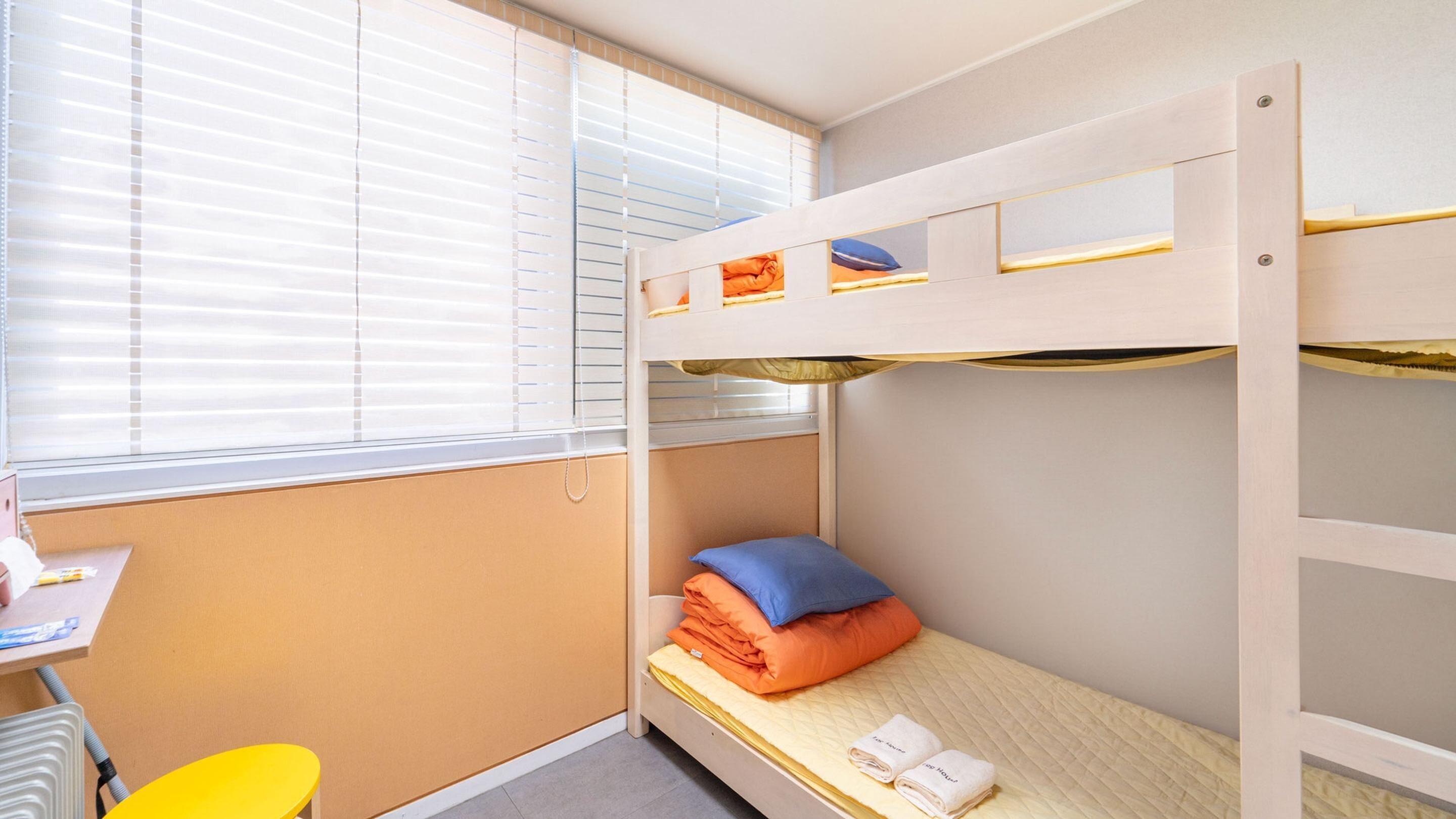 A bunk bed room with a desk and chair, a window with blinds, and a small table with a chair.
