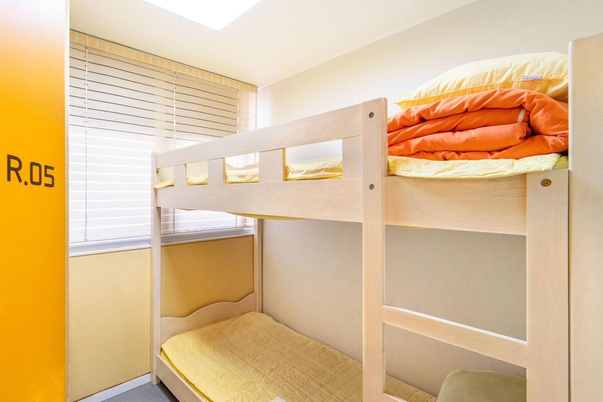 Une chambre avec un lit superposé, une fenêtre et des stores, un lit superposé avec une literie orange, et un mur sur lequel on peut lire le texte « EGG R.05 ».