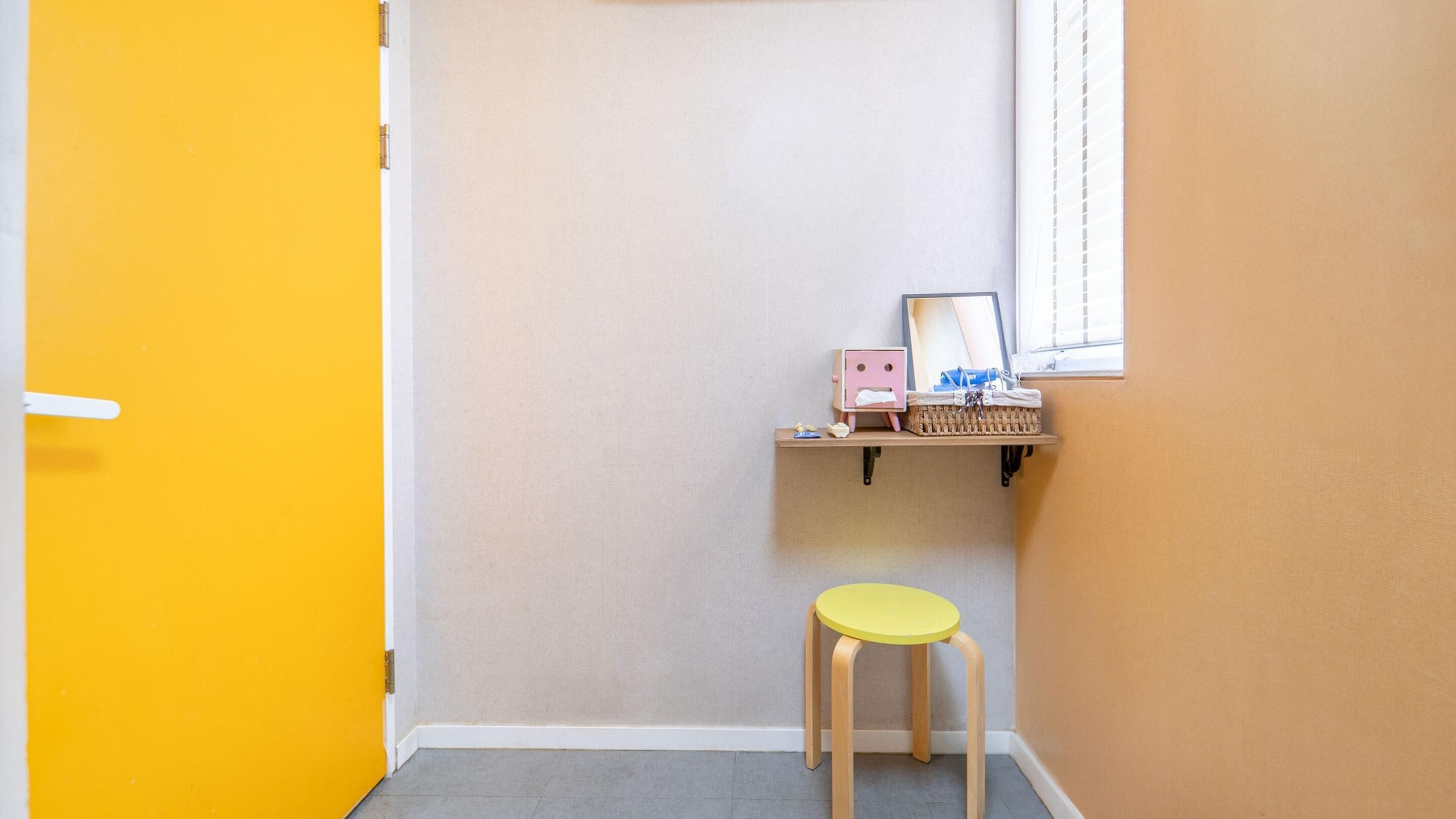 Une pièce d’angle avec une porte jaune, un petit tabouret, une étagère avec un panier et une fenêtre.