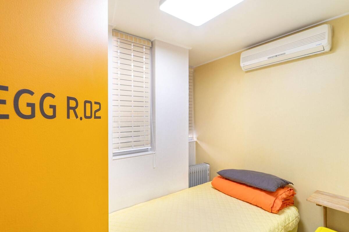 Une pièce avec un mur jaune portant l’inscription « EGG R.02 », une fenêtre avec des stores, un lit avec des oreillers et un climatiseur fixé au mur.