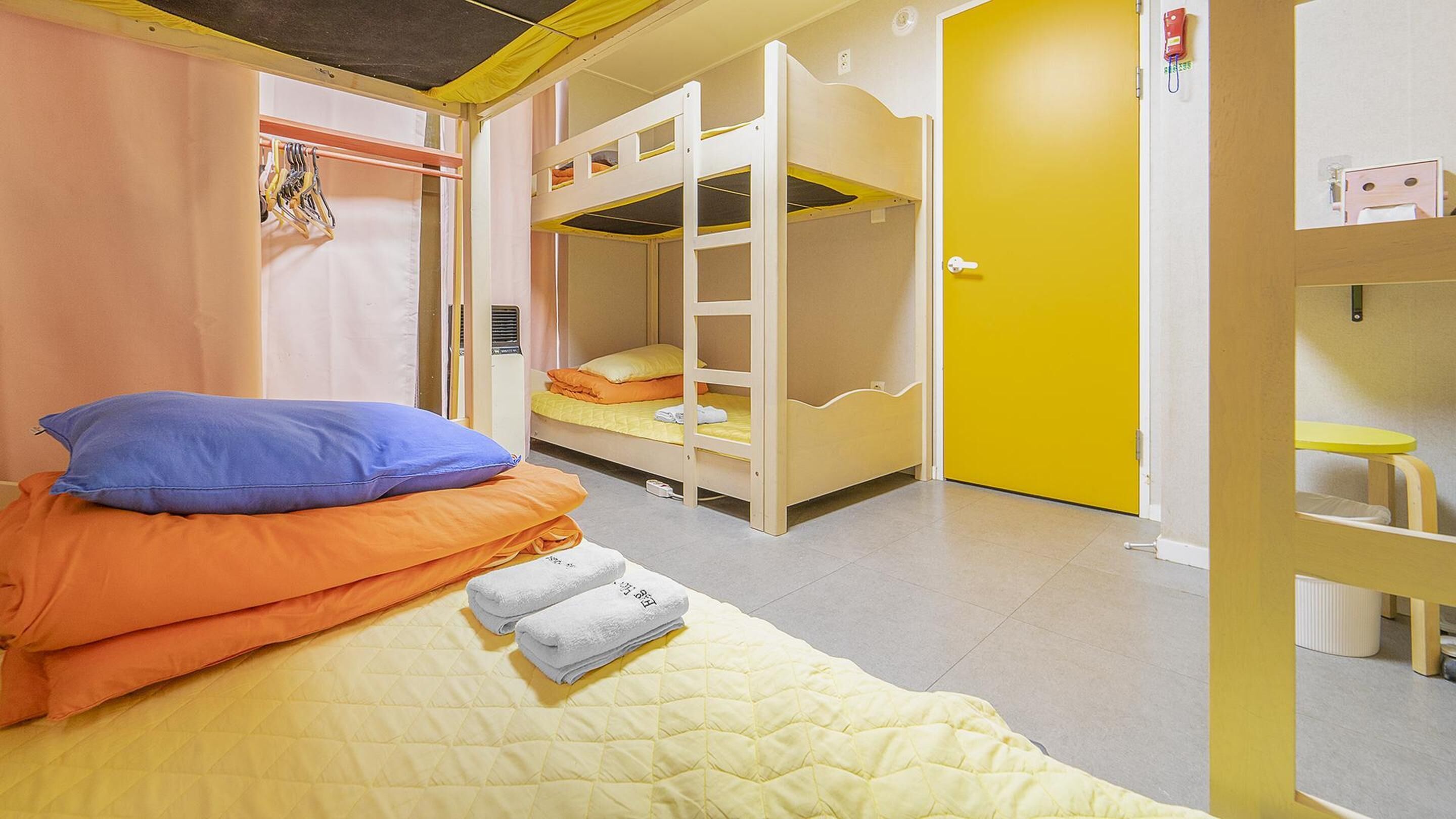 Una habitación con literas, una cama con almohadas, una puerta amarilla y una pequeña mesa.