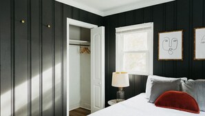 Ein Schlafzimmer mit einem Bett, einem Nachttisch, einer Lampe, einem Fenster mit Kunstwerk und einem Kleiderschrank.