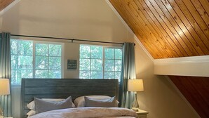 Une chambre avec un plafond en pente, un lit doté d’une tête de lit en bois, deux lampes de chevet et une fenêtre avec des rideaux.