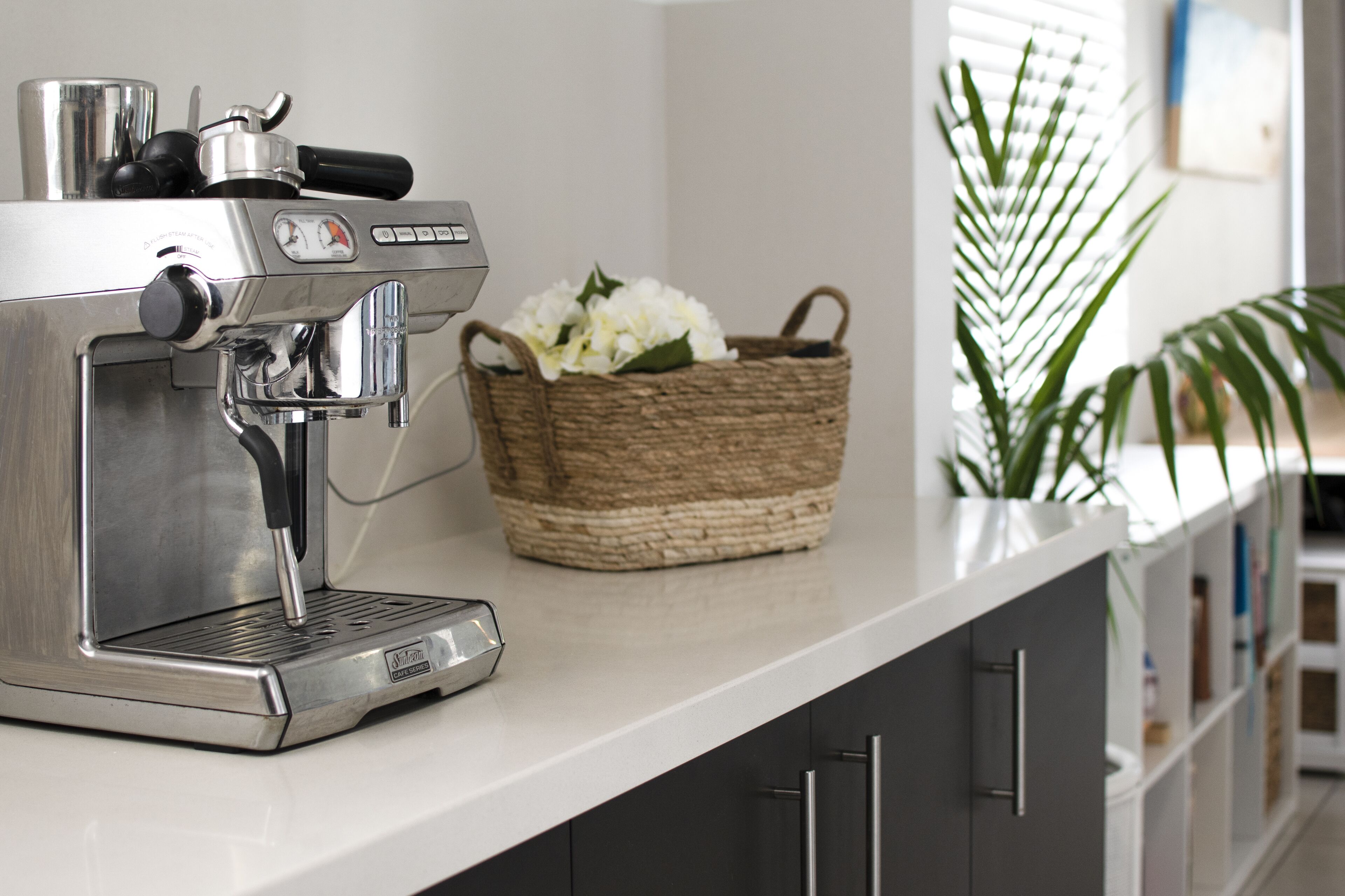 A kitchen with a stainless steel espresso machine, a woven basket with white flowers, and a potted plant.