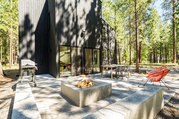 Relax in the serene patio area with a fire pit, seating, and a grill, surrounded by tall trees.