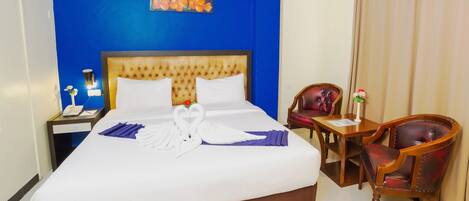 A hotel room with a large bed, a wooden headboard, a desk with a chair, and a blue wall with a floral painting.