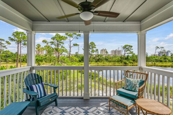 A covered porch with two Adirondack chairs, a ceiling fan, and a view of a green landscape with trees and buildings in the distance.