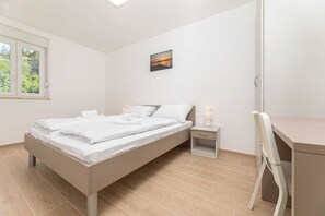 Un dormitorio con cama, mesita de noche, silla, escritorio y un cuadro en la pared.
