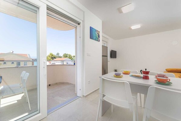 Un balcon avec une porte coulissante, une table à manger avec des chaises, un téléviseur fixé au mur et un petit coin cuisine.