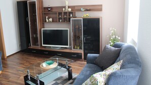 A living room with a flat-screen TV, wooden shelving with decorative items, a glass coffee table, and a grey sofa. - Modernized apartment, centrally located in historic old town (Mücheln (Geiseltal))