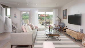 Amplia sala de estar con sofá, mesa de centro y televisor. Se ve un balcón con mobiliario exterior a través de una puerta de cristal.