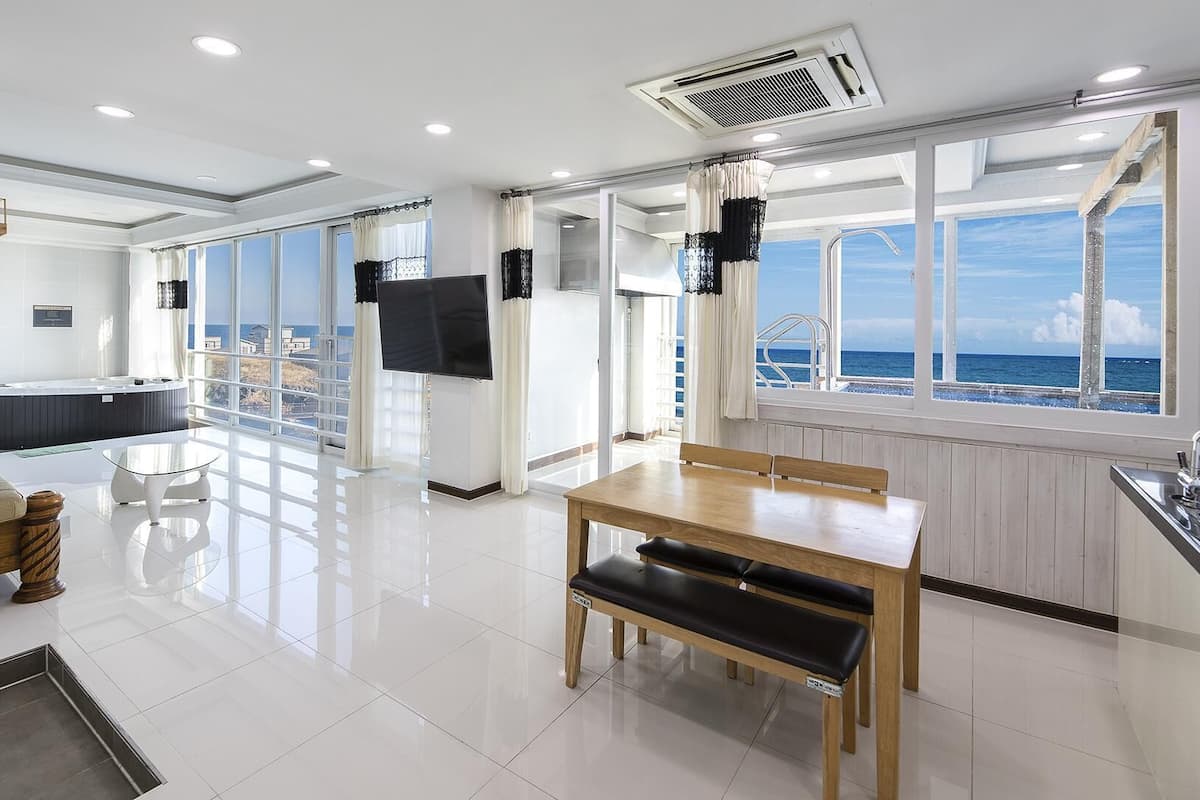 A modern, well-lit room with a dining area, a sofa, and a view of the sea through large windows.
