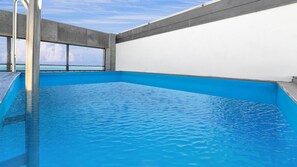 An indoor swimming pool with clear blue water, a metal ladder, and a view of the sea through large windows.