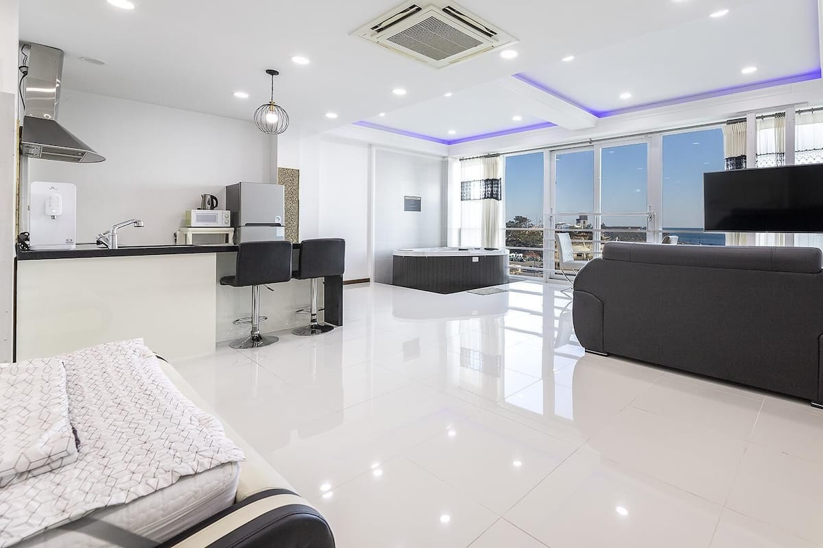 A modern living room with a sofa, a coffee table, and a TV. There is a balcony with a view of the city.
