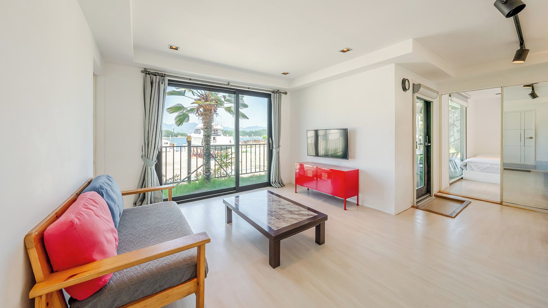 Un moderno salón con un sofá, una mesa de centro, un televisor y un balcón con vistas a palmeras y un paisaje lejano.