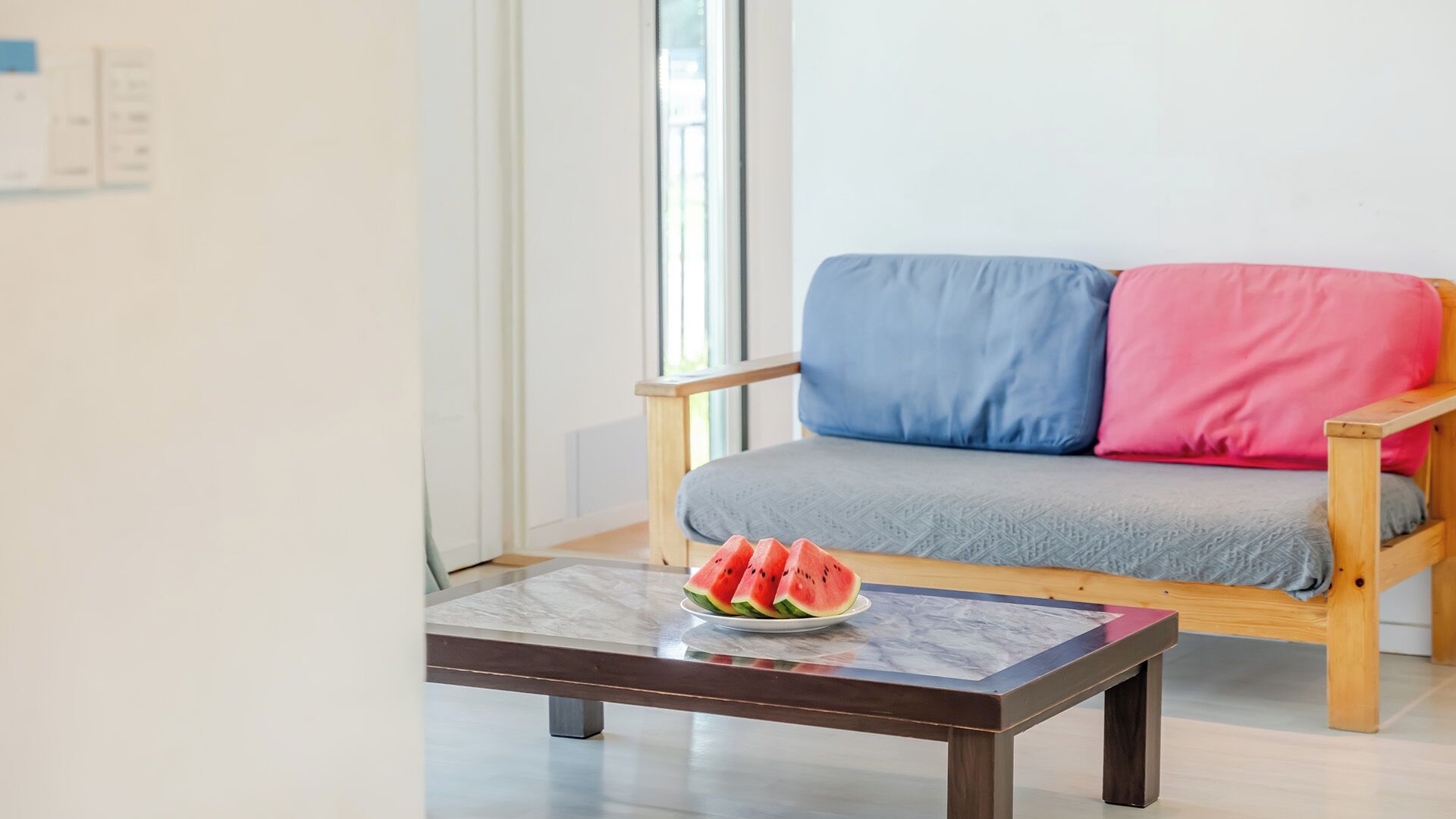 Una habitación con un sofá de madera, una mesita con una fuente de sandía en rodajas y una ventana.