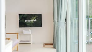 Un salón moderno con un televisor de pantalla plana montado en la pared, una mesita blanca, un banco de madera y vistas a un balcón a través de cortinas translúcidas.