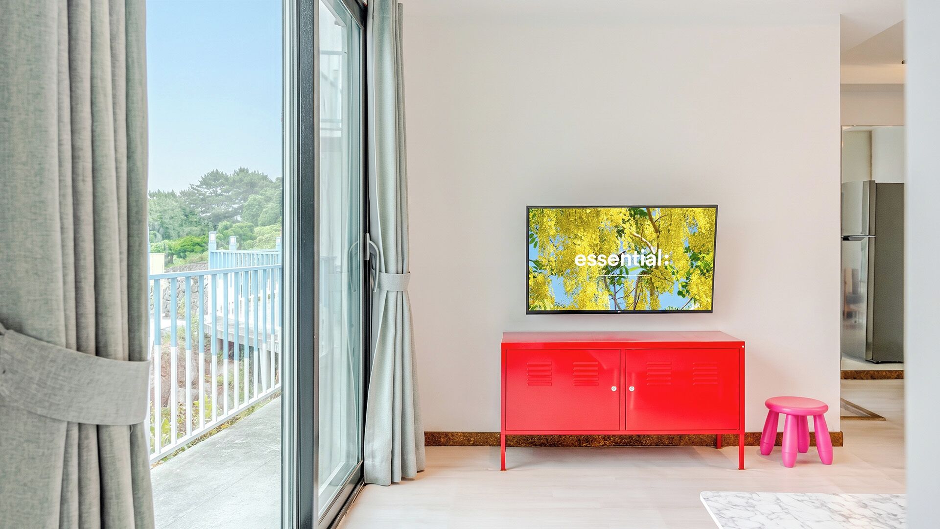 A modern living room with a large flat-screen TV mounted on a white wall, a red cabinet below it, and a pink stool to the right.
