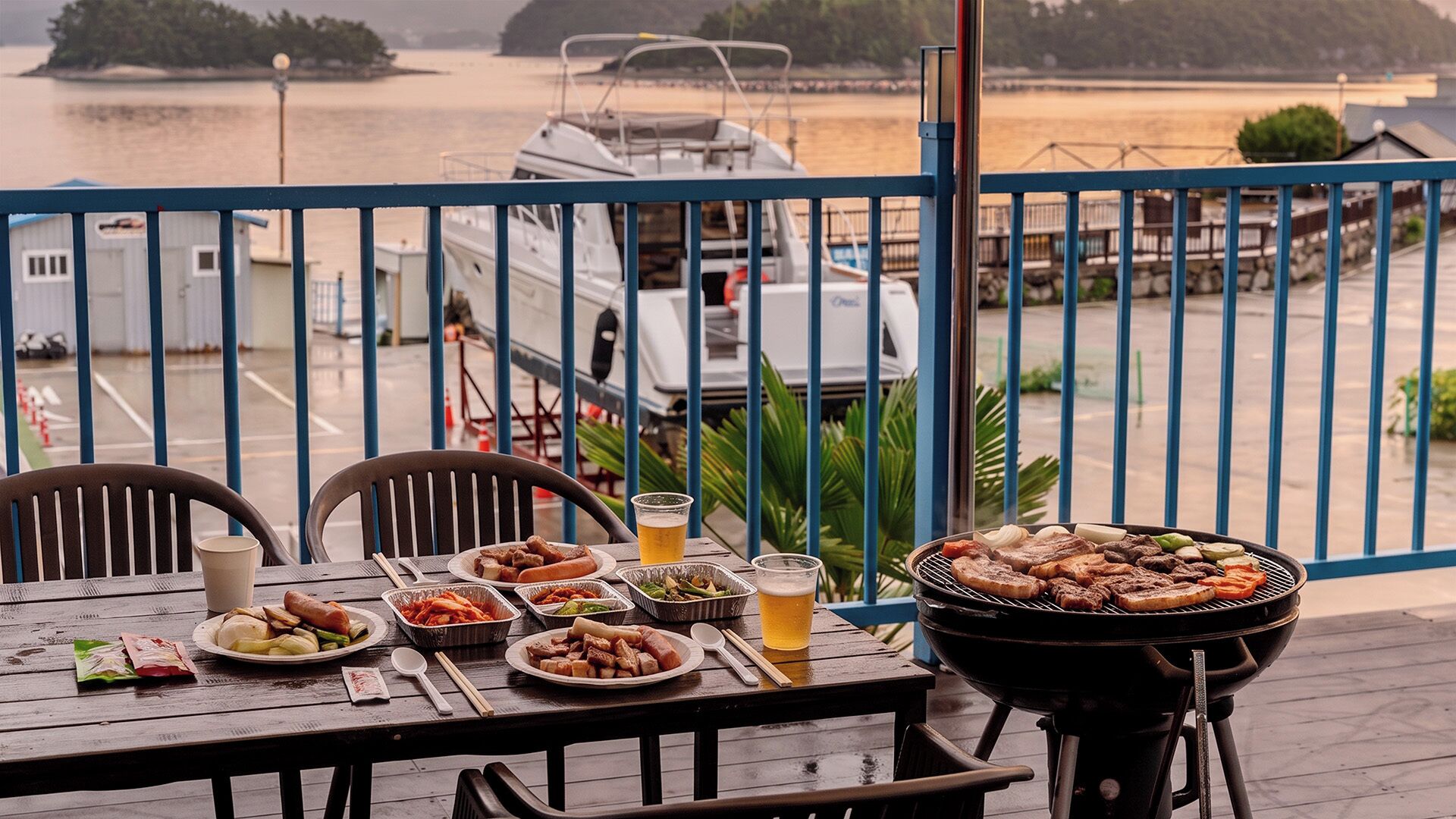 Una mesa de comedor con diversos platos y bebidas, una parrilla, y vista a barcos y al agua.