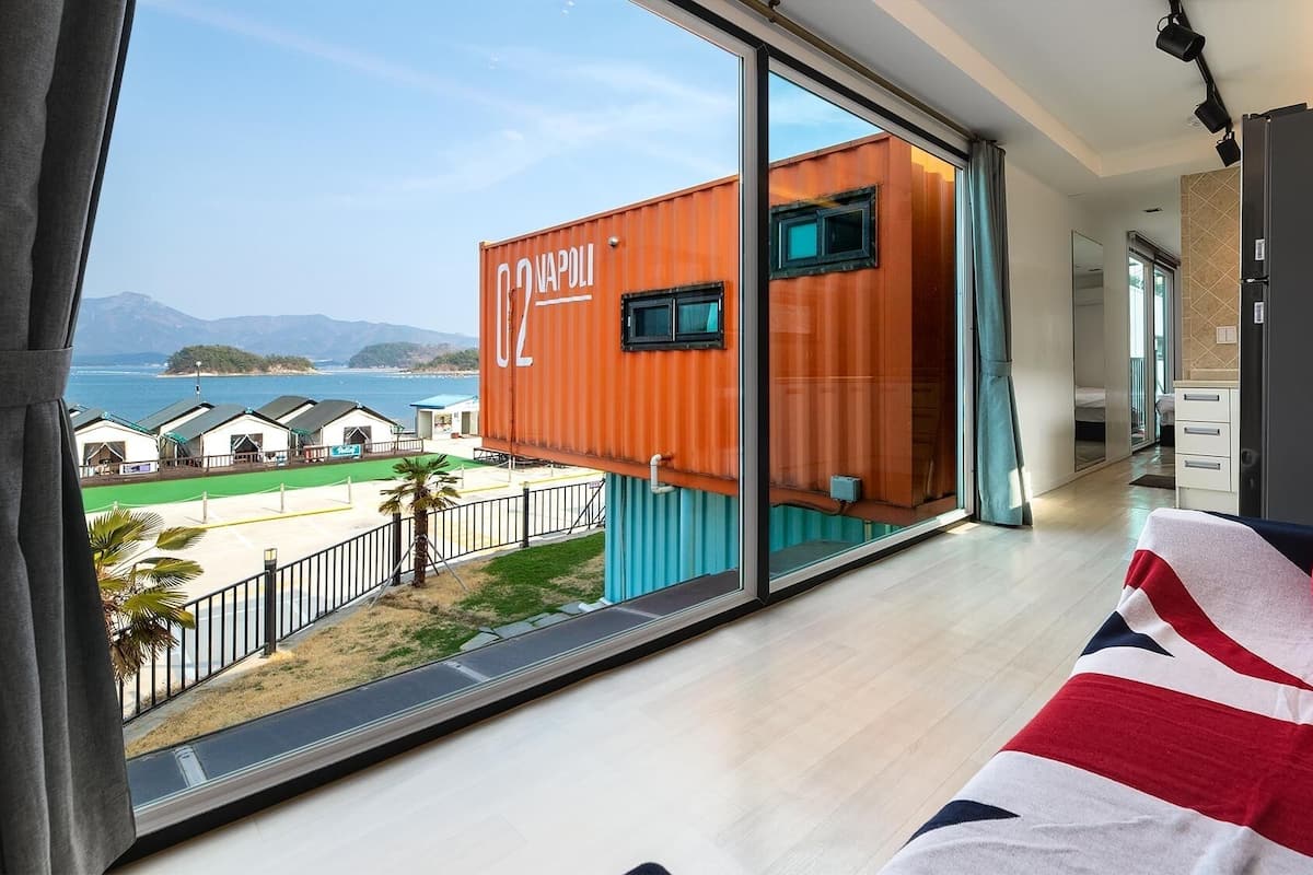 Une chambre moderne dotée d’une grande fenêtre, d’un canapé et offrant une vue sur une zone côtière où se trouvent des conteneurs de transport.