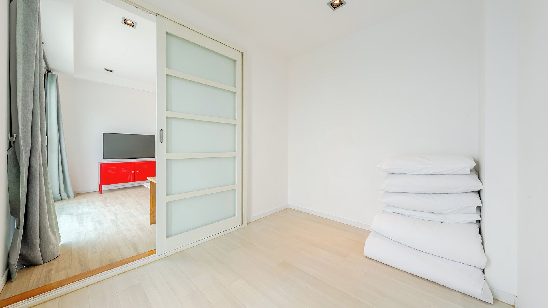 Una habitación con puerta corredera, un armario rojo, un televisor y una pila de almohadas blancas.