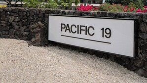 Une enseigne indiquant « PACIFIC 19 », avec un bâtiment en arrière-plan, des palmiers et un mur de pierre.