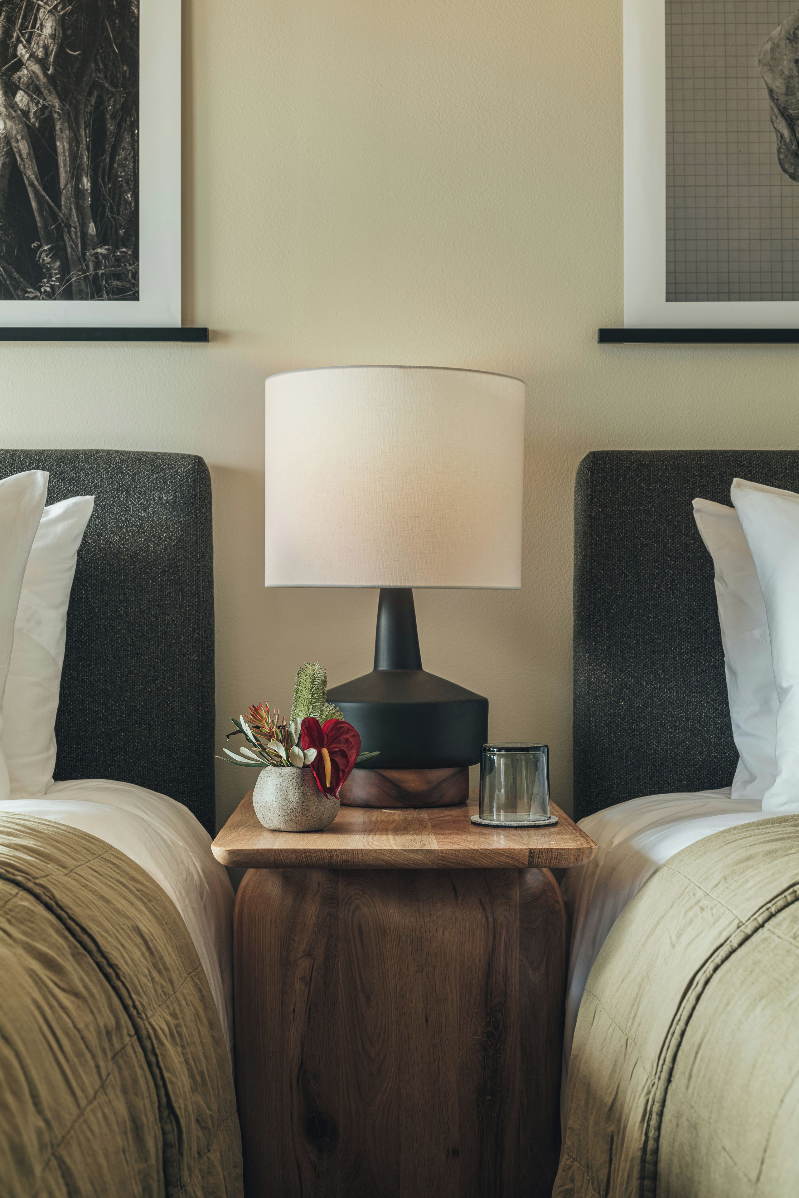 A hotel room with two beds, a bedside table with a lamp, a vase with flowers, and a glass of water.