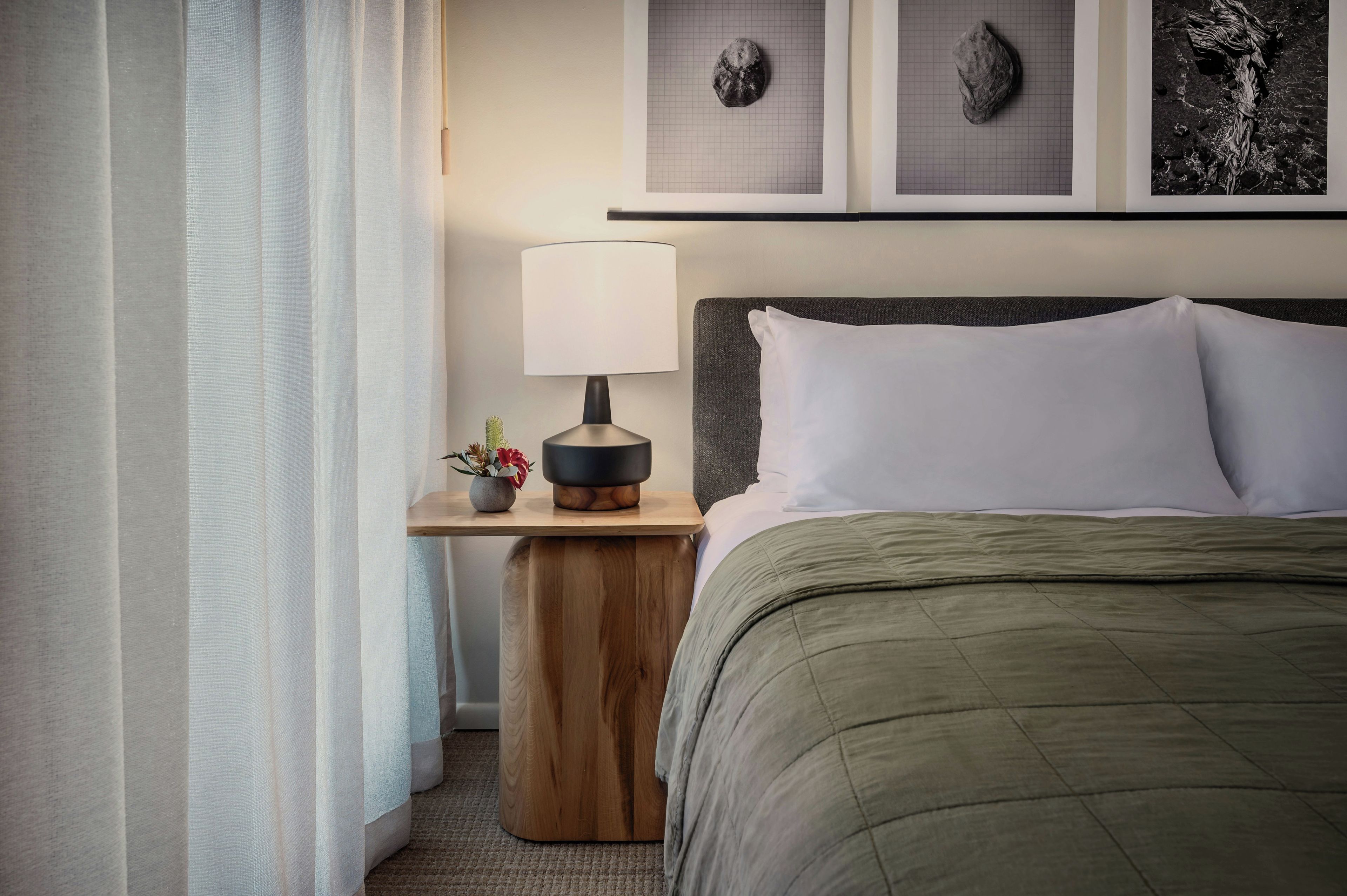 A neatly made hotel room with a bed, a bedside table with a lamp, a small plant, and artwork on the wall.