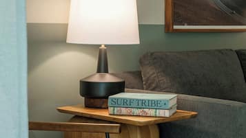 A room with a lamp on a wooden side table, a book titled 'SURF TRIBE' on the table, a couch, and a painting on the wall.